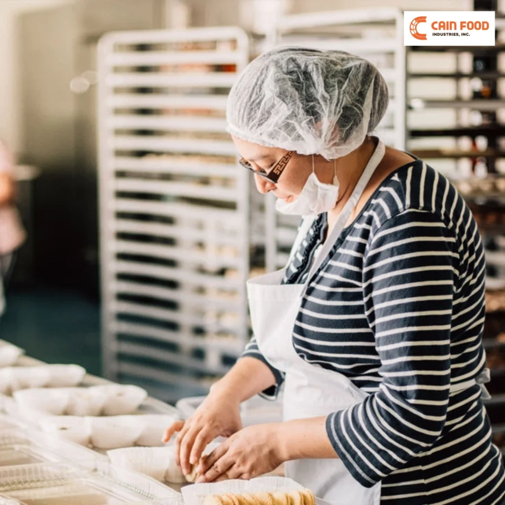 Baker in a striped shirt and white apron shaping dough at a bakery station, wearing a hairnet and mask.