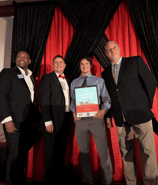 Four men in formal attire on a stage, with one holding a framed award certificate against red curtains in the background.