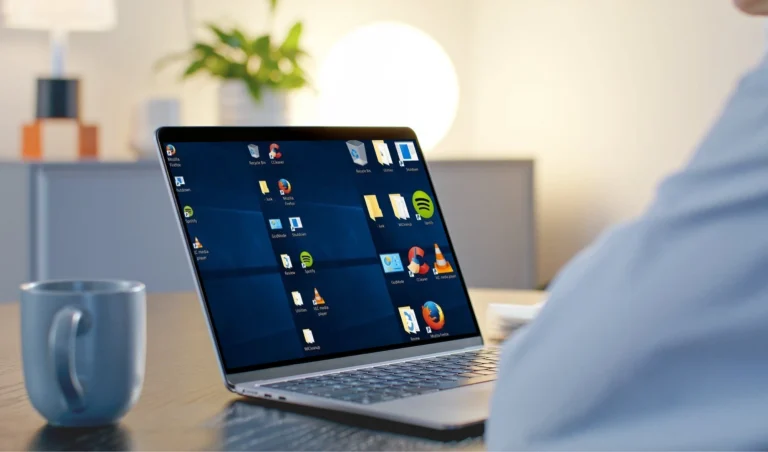 Laptop on a desk with its screen showing a grid of app icons, a mug in the foreground and a blurred office background.