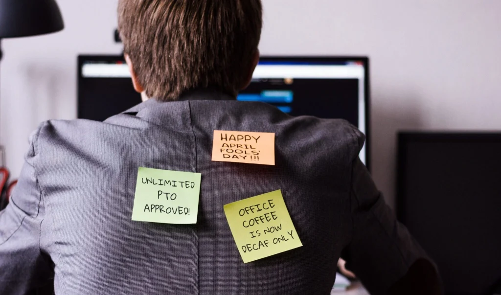 Person seen from behind with colorful sticky notes on a blazer containing humorous workplace messages (Happy April Fools' Day!).