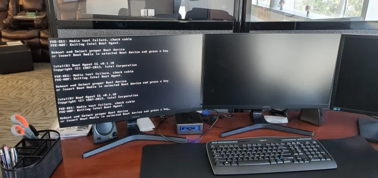 Triple-monitor computer workstation with a left monitor showing repeated BIOS/boot error text (PXE-E61), middle and right screens off; keyboard and mouse on a wooden desk.