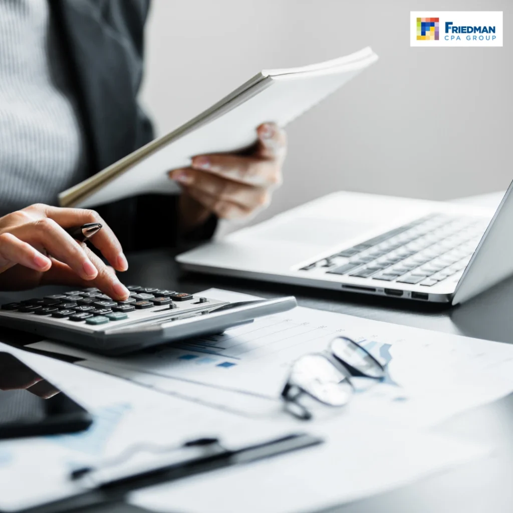 Businessperson calculating numbers with a calculator beside a laptop and financial documents; Friedman CPA Group logo visible.