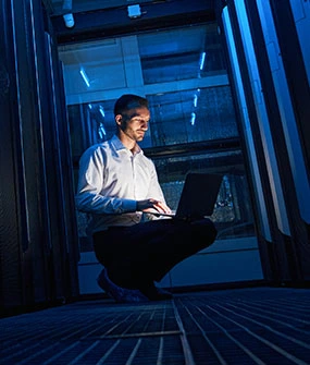 Man kneeling in a data center, typing on a laptop beside blue-lit server racks.