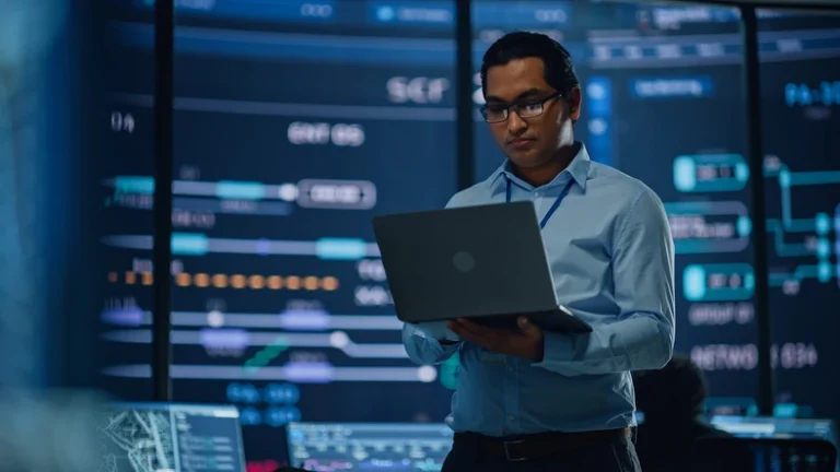 Man in a light blue shirt and glasses reviews data on a laptop in a high-tech control room with multiple screens displaying graphs and charts.