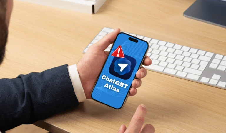 Man in a suit holding a smartphone showing a ChatGPT Atlas app with a red warning triangle over the screen, on a wooden desk with a keyboard in the background.