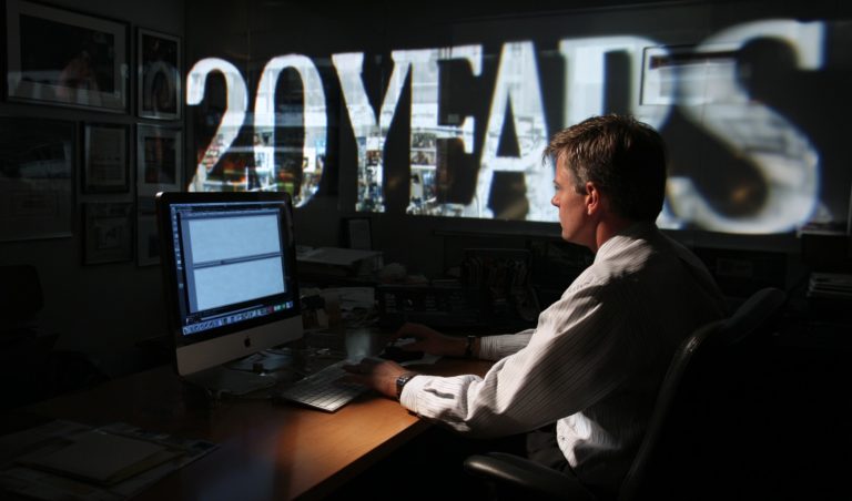 Man in a striped shirt sits at a desk typing on a keyboard, with a large wall projection behind him showing graphics.