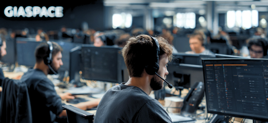 Open-plan office with multiple staff wearing headsets and working at computer desks in a busy tech environment (GIASPACE branding in background).
