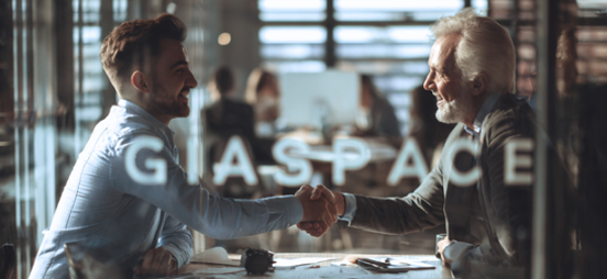 Two men shake hands across a conference table in a modern office, seen through a glass wall with a GLASPACE sign visible in the foreground a collaboration moment.