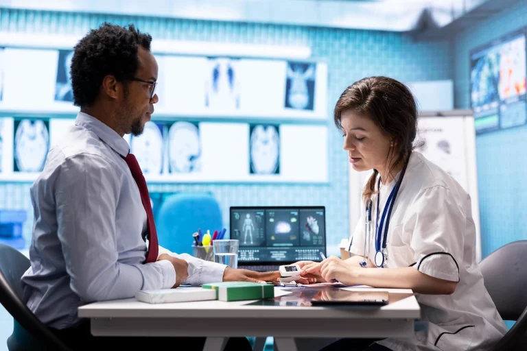 Doctor with stethoscope meets a patient at a clinic desk; a laptop and medical images are visible on screens in the background.