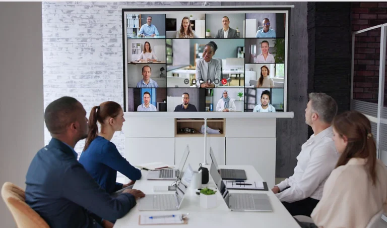 Team in a meeting room participates in a video conference shown on a large display with multiple remote participants.