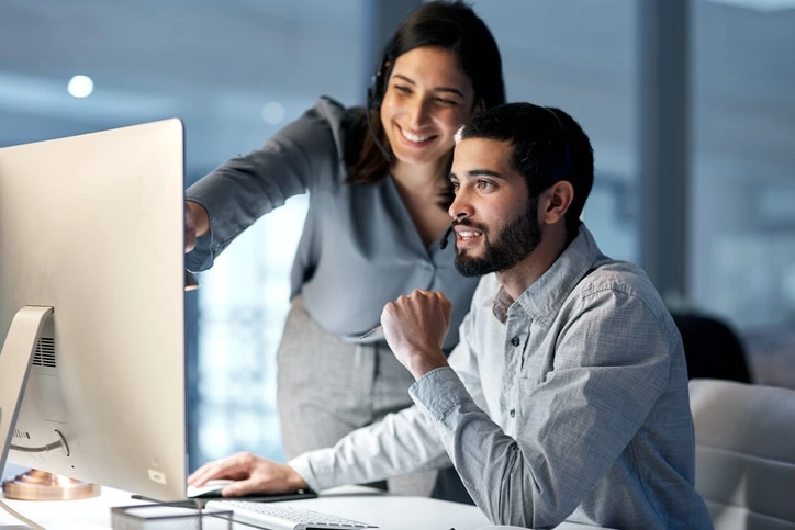 Two coworkers work at a desktop computer; woman points at the screen while smiling.