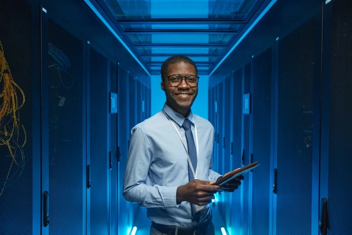 IT professional in a server room, holding a tablet and smiling at the camera under blue neon lights.