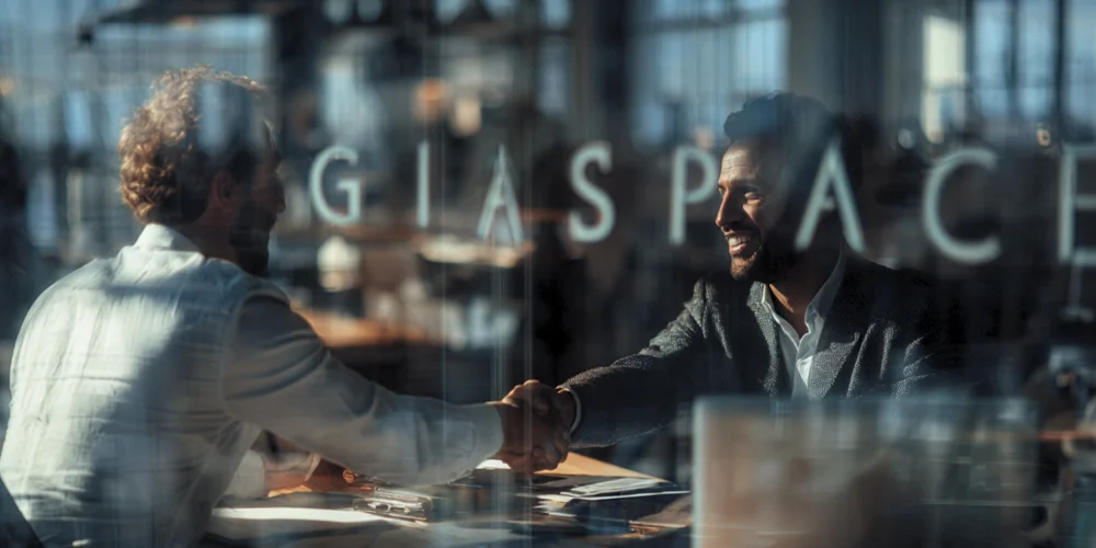 Two businessmen shake hands across a desk in a glass-walled office.