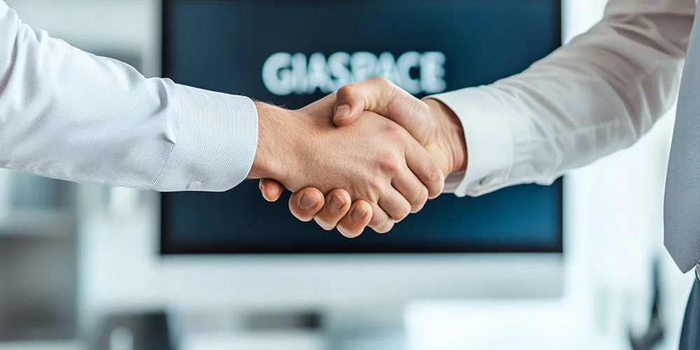 Two professionals in an office shake hands, signaling a business agreement or partnership in front of a blurred screen in the background.