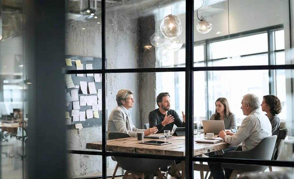 Five professionals sit around a wooden table in a glass-walled meeting room, discussing with laptops and sticky-note wall in the background.